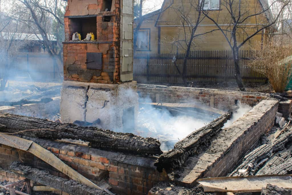 A brick chimney and some walls remain standing amid the smoldering ruins of a burned-down house, with charred wooden beams and smoke rising from the debris. A wooden fence and house are visible in the background.