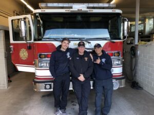 Three firefighters in uniform stand smiling in front of a red and white fire truck inside a fire station garage. Two have their hands on their hips, while one gestures with a thumbs-up.