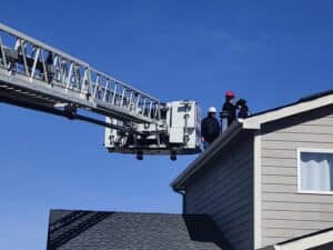 Three people wearing helmets and safety gear stand on the roof of a house near a raised fire truck ladder under a clear blue sky.
