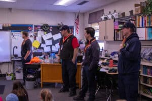 Four firefighters stand at the front of a classroom, facing seated students. The classroom has a cluttered desk, shelves with books, and a bulletin board with papers. An American flag hangs by the window.