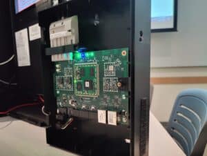 A close-up view of an open electronic device casing reveals a green circuit board with various components, glowing indicator lights, and connected wires, set on a desk in a classroom or office environment.