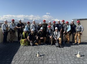 A group of 18 people wearing safety gear, helmets, and harnesses pose together on a rooftop with mountains and a blue sky in the background. Some hold ropes and equipment.