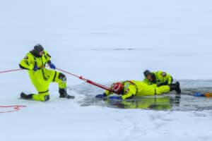 Three rescue workers in bright yellow suits perform an ice rescue drill; one worker pulls on a rope while another reaches into a hole in the ice, and a third assists on the snowy surface.