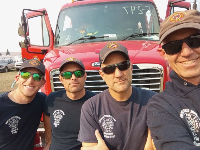 Four firefighters in navy shirts and caps pose smiling in front of a bright red fire truck on a sunny day.