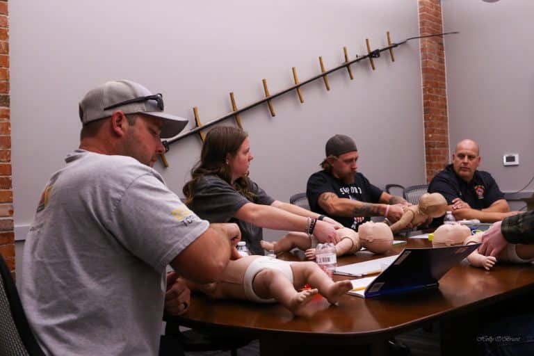 Four adults sit around a conference table practicing infant CPR techniques on baby mannequins; training materials and laptops are on the table in a room with brick walls and a large stick hung on the wall.