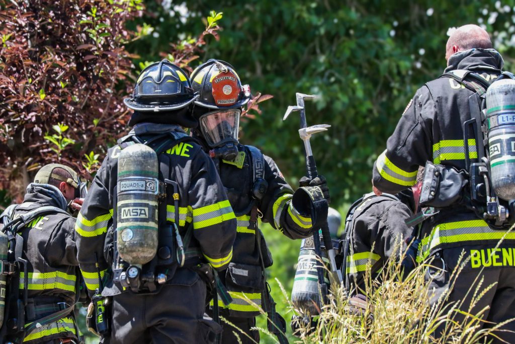 A group of firefighters in full gear, including helmets and oxygen tanks, standing together outdoors. One firefighter in the center holds a halligan tool, while others face away, surrounded by greenery and tall grass.